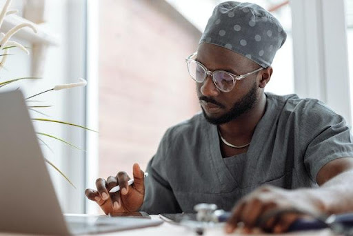 A black male doctor wearing gray scrubs working on a laptop.
