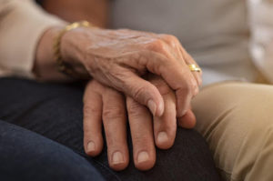 Closeup of an elderly woman holding the hand of an elderly man on her knee.