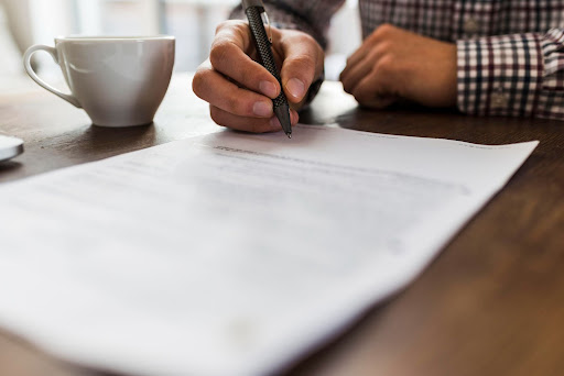 a man in a checkered shirt sitting at the table with a cup of coffee signing a document