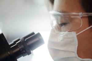A man wearing glasses and a mask looking through a microscope