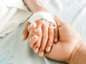 Close-up of a bed-ridden hospital patient’s hand enclosed in the hand of a loved one